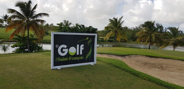 A golf course sign with a logo and the name 'Golf de Saint-Francois' on a grassy area with palm trees and a body of water in the background. - Touba Manga Beach (Saint-François)