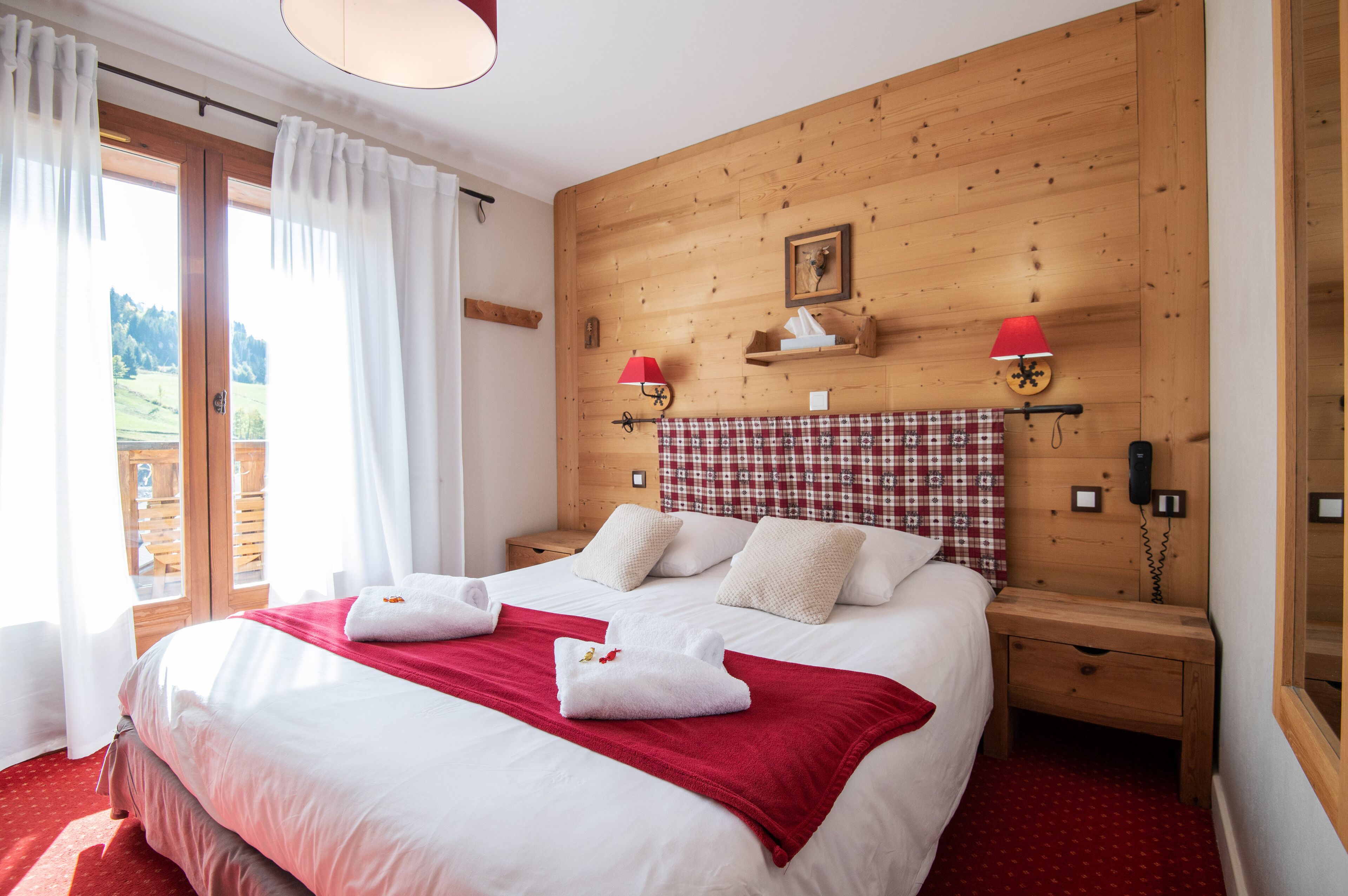 A hotel room with a large bed, wooden headboard, a red checkered curtain, and a wooden nightstand with a lamp.