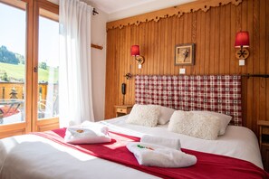 A hotel room with a wooden paneled wall, a checkered headboard, a bed with white bedding and red accents, and a view of a balcony with outdoor furniture. - Hôtel L'Alpage (Le Grand-Bornand)