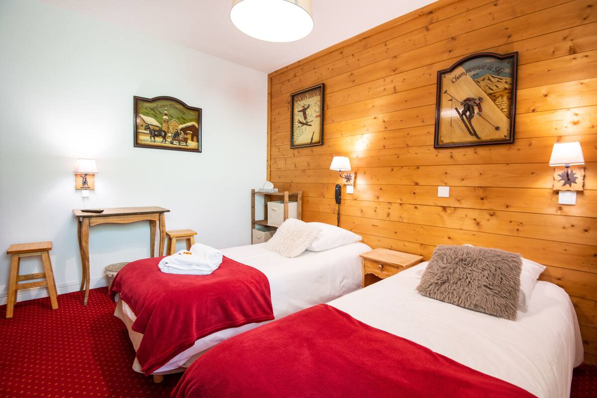 A hotel room with two beds, wooden walls, and a red carpet. There are two bedside tables with lamps and a small desk with a chair.