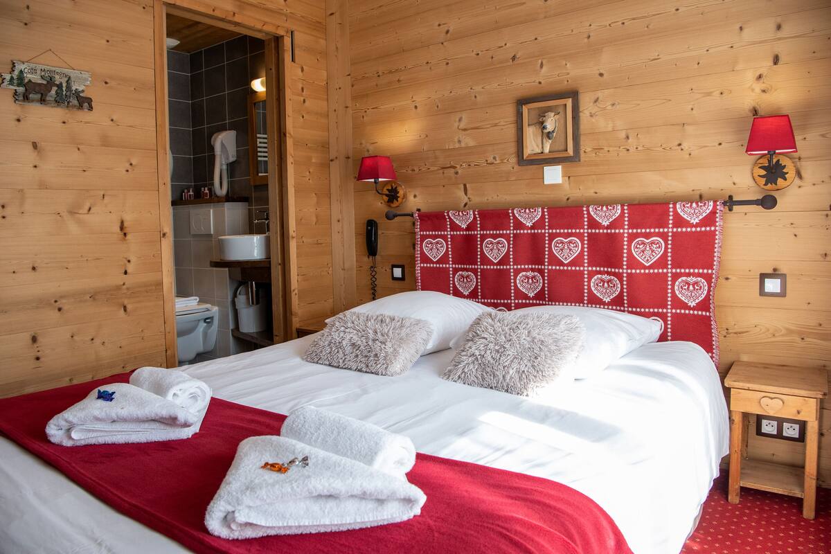A wooden-paneled room with a bed, towels, a red headboard, a small bedside table, and a wall-mounted lamp.