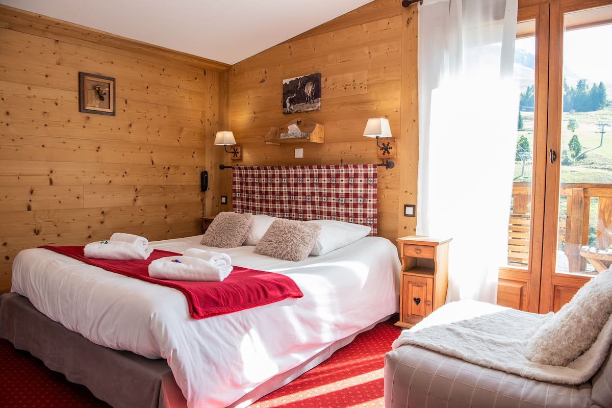 A wooden-paneled room with a bed, two bedside lamps, a red and white checkered headboard, and a view of a mountainous landscape through a glass door.