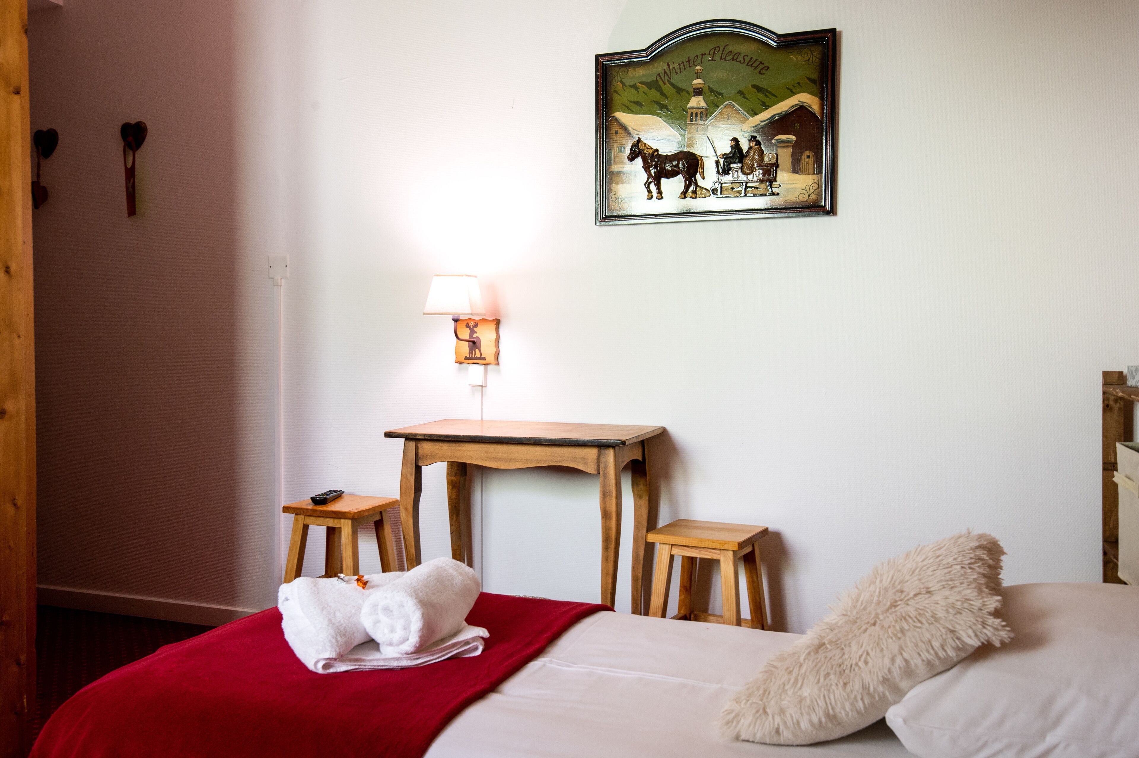 A neatly arranged hotel room with a bed, a small wooden table, a wall-mounted lamp, and a painting depicting a winter scene.