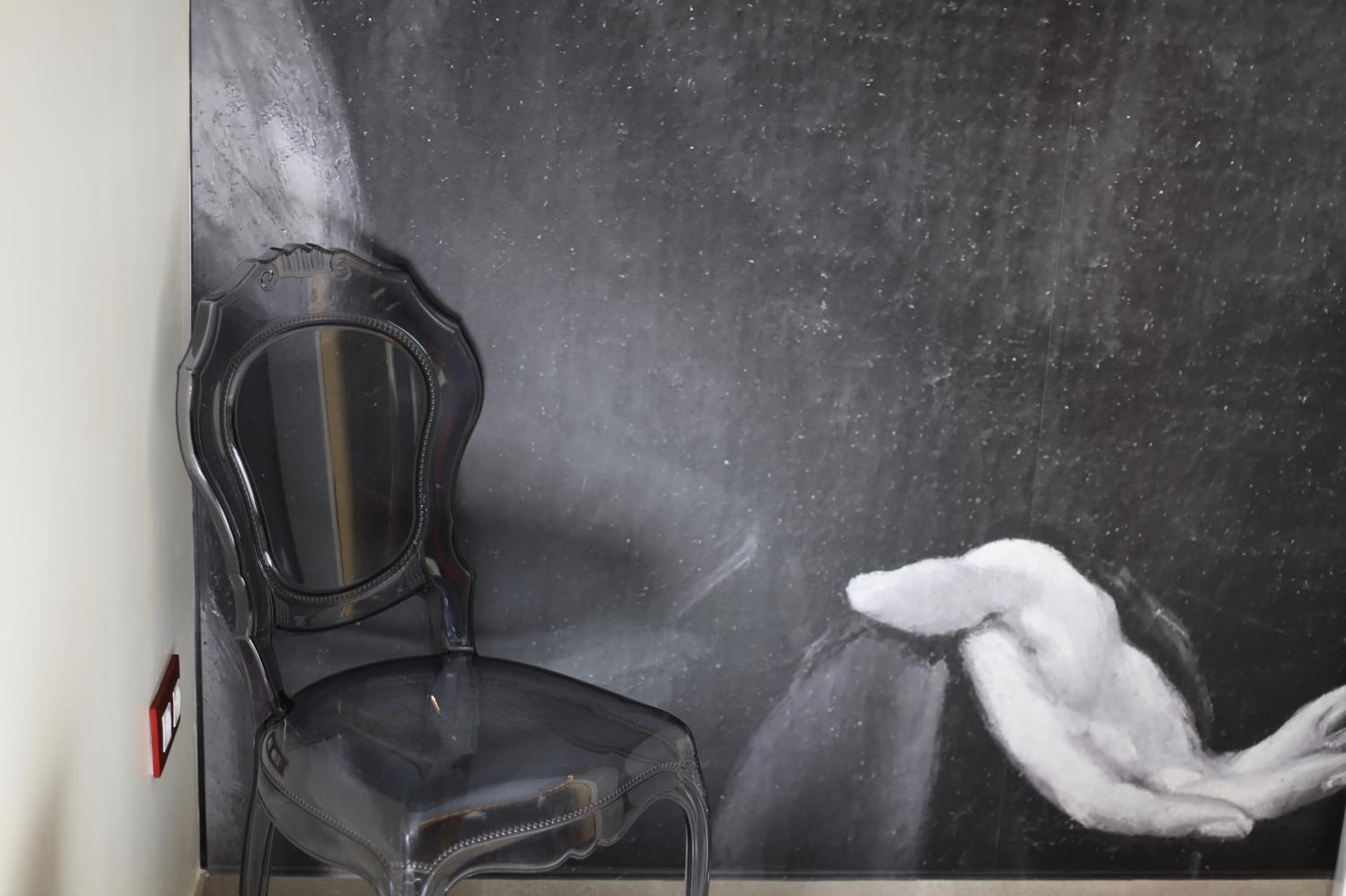 Een stoel met een transparante zitting en rugleuning, geplaatst tegen een muur met een schilderij van een witte figuur die reikend is.