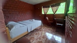 A room with a brick wall, a bed with a white bedspread, a wooden bench, and a window with curtains.