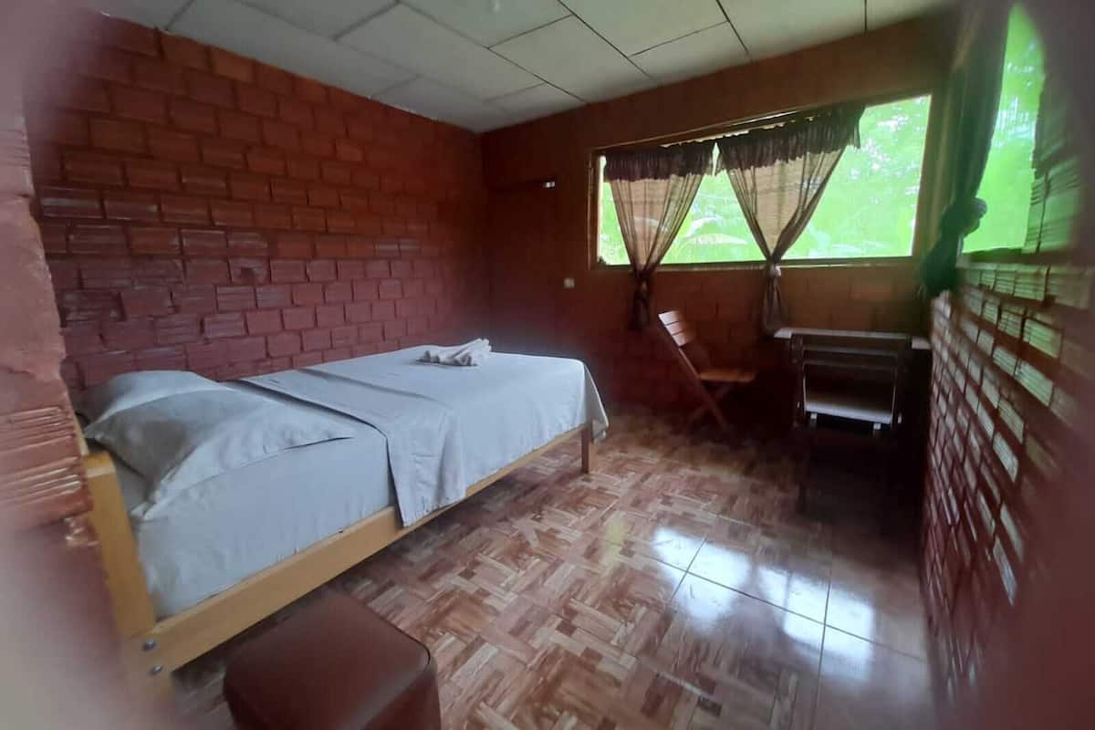 A room with a brick wall, a bed with a white bedspread, a wooden bench, and a window with curtains.