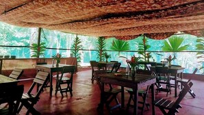 An outdoor dining area with wooden tables and chairs, surrounded by potted plants and a ceiling with a floral pattern.