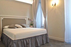 A neatly made bed with a headboard, a bedside table with a vase of flowers, and a wall-mounted light fixture. - Locanda San Tomaso (Pietra Ligure)