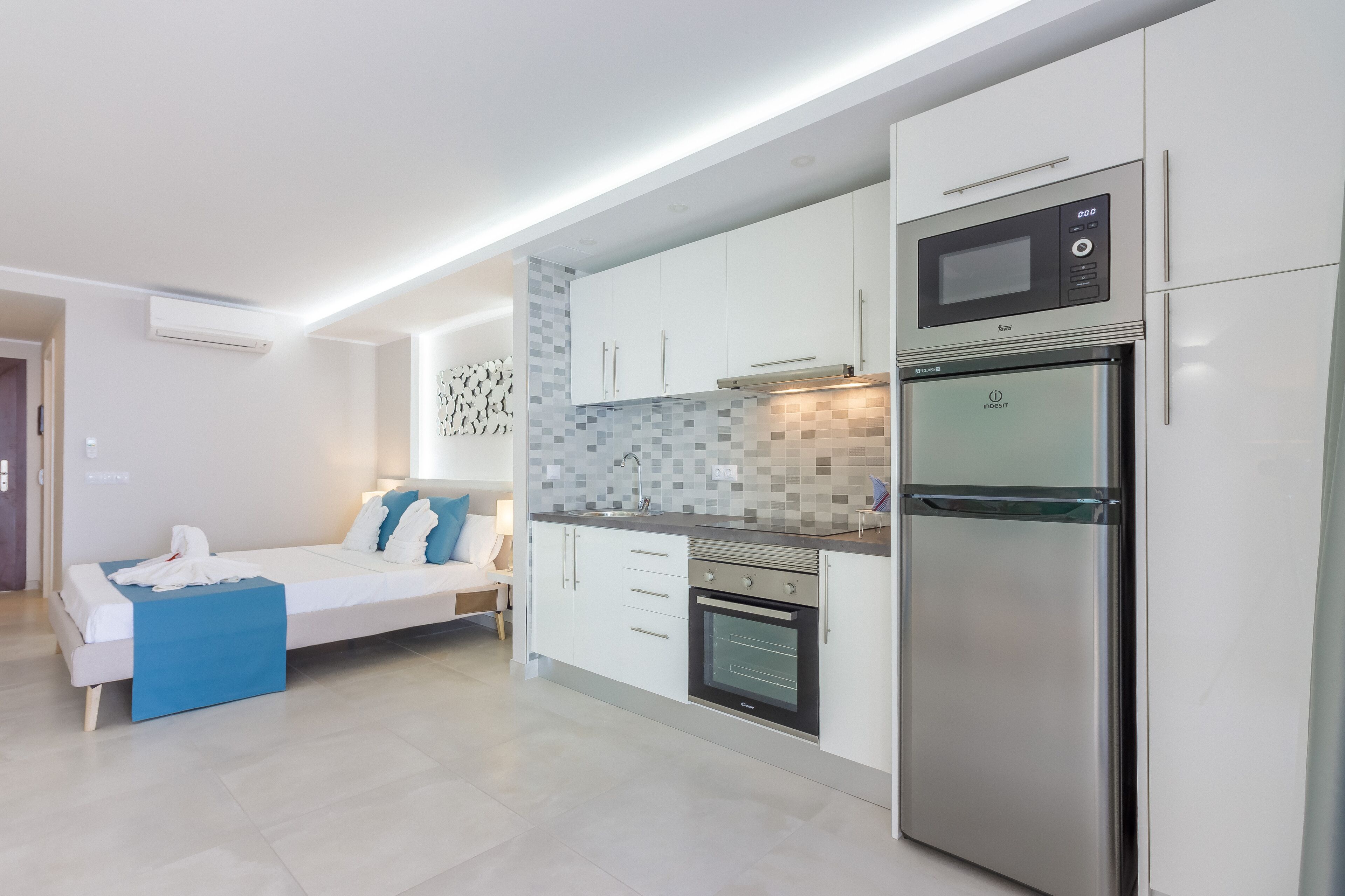 A modern kitchen with a built-in microwave, oven, and refrigerator. There is a bed with a blue throw blanket and pillows in the adjacent room.