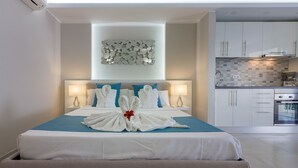 A hotel room with a neatly made bed, a modern kitchen in the background, and a decorative wall piece above the bed.