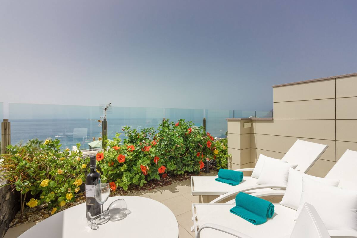Eine Terrasse mit weißen Liegestühlen, einem Tisch mit einer Weinflasche und einem Glas sowie Blick aufs Meer.