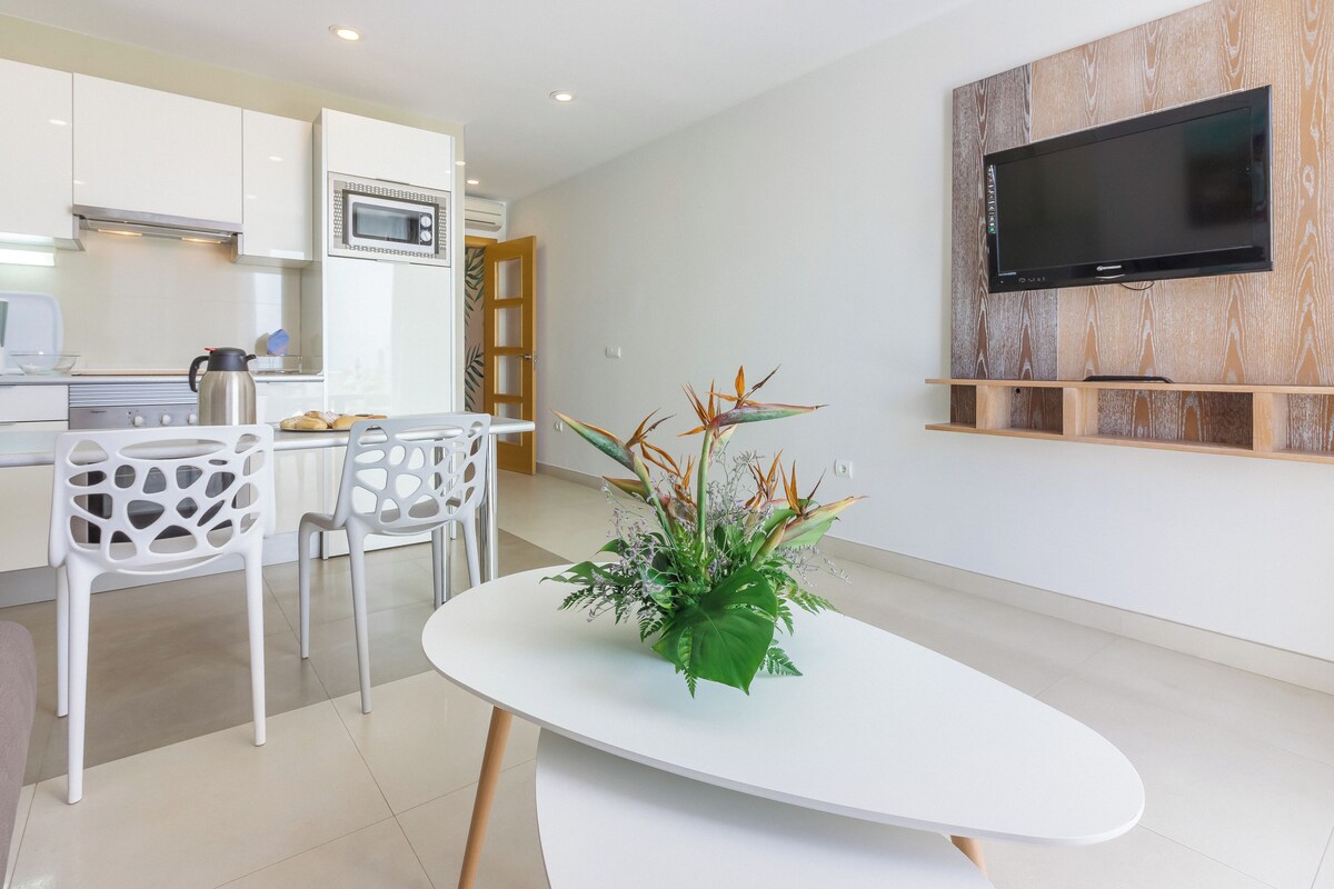 A modern kitchen with white cabinets, a microwave, and a coffee maker. There is a dining area with a white table and chairs. A flat-screen TV is mounted on the wall.