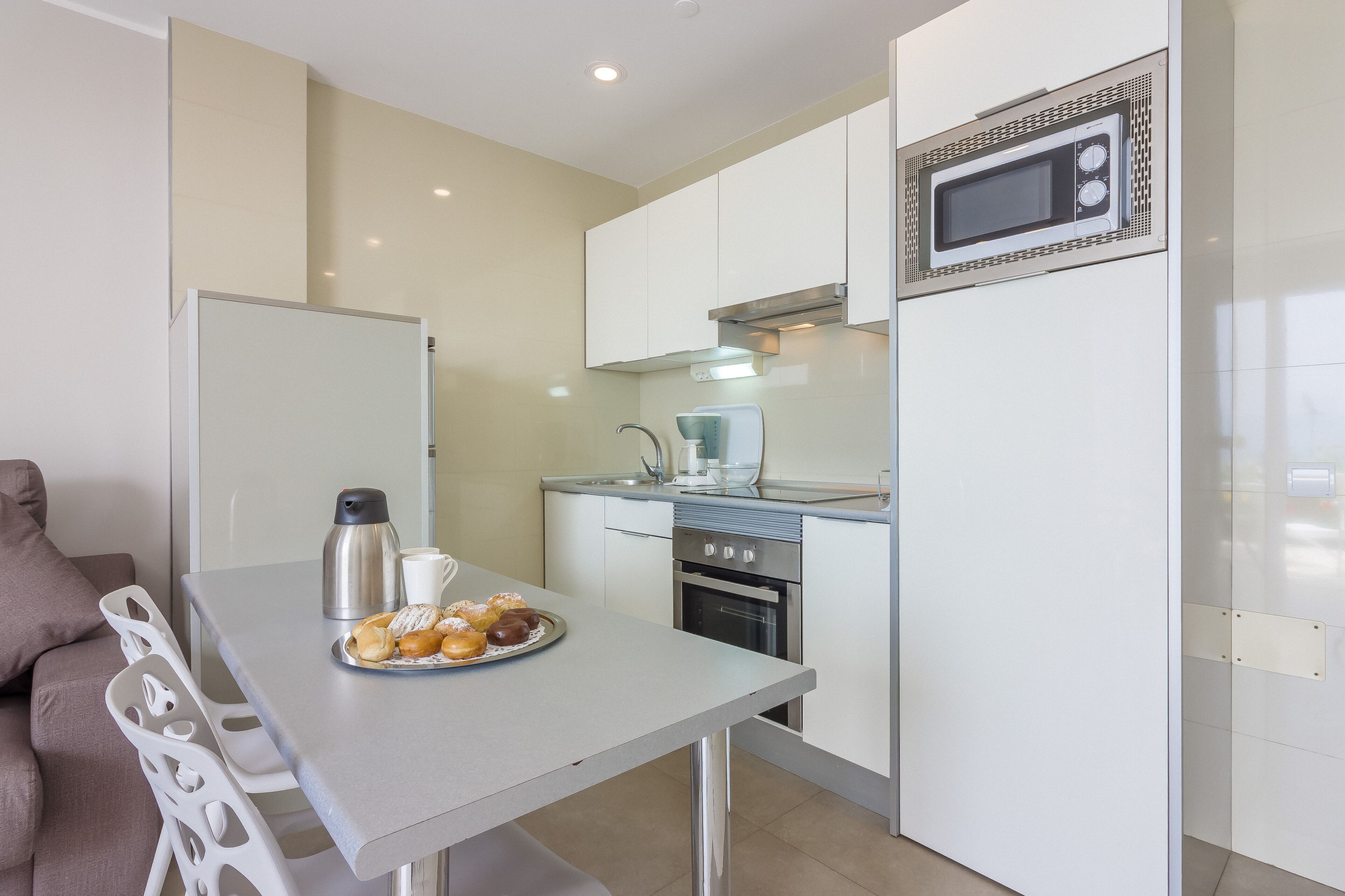A compact kitchen with a dining area, a microwave, a kettle, and a plate of pastries.