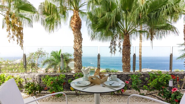 A table set outdoors with a view of the ocean, surrounded by palm trees and a stone wall.