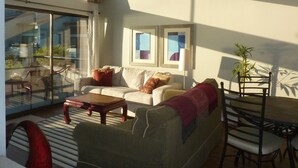 Ein Wohnzimmer mit einer Couch, einem Couchtisch und einem Esstisch mit Stühlen. Ein Balkon bietet Blick ins Freie.