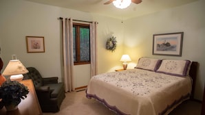 Un dormitorio con cama, una silla, una lámpara, una ventana con cortinas, un arreglo en la pared y un cuadro enmarcado.