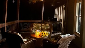 Une terrasse en bois agrémentée de jardinières suspendues et de guirlandes lumineuses, un banc rembourré avec une lanterne, et une vue sur l’intérieur à travers une fenêtre.