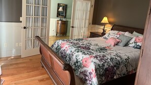 Une chambre à coucher avec un lit en bois, une literie à motifs floraux, une table de chevet avec une lampe, une commode et une porte donnant sur une autre pièce.