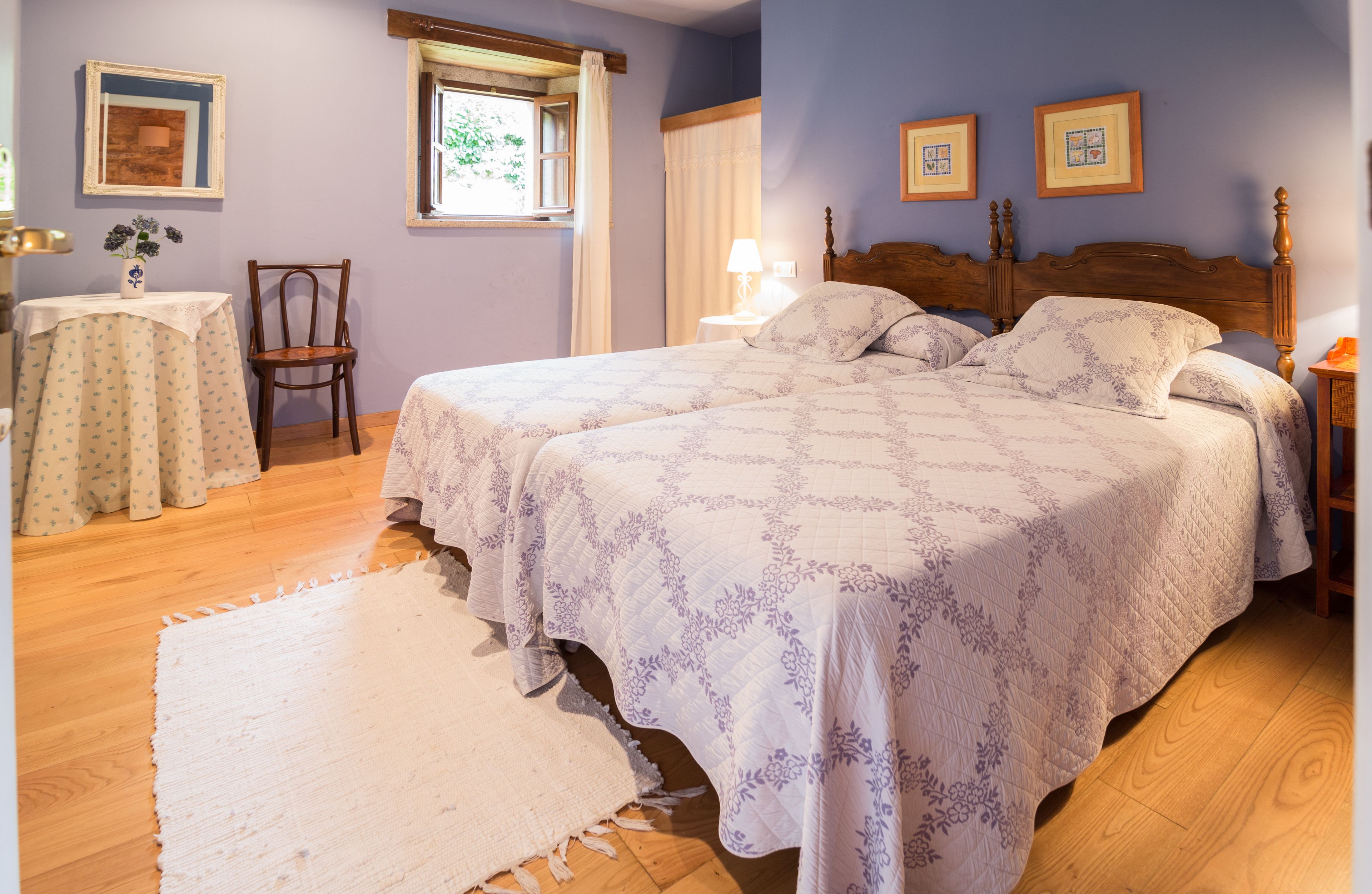 Quarto com duas camas, piso de madeira, uma mesinha com um vaso e uma janela com cortinas.