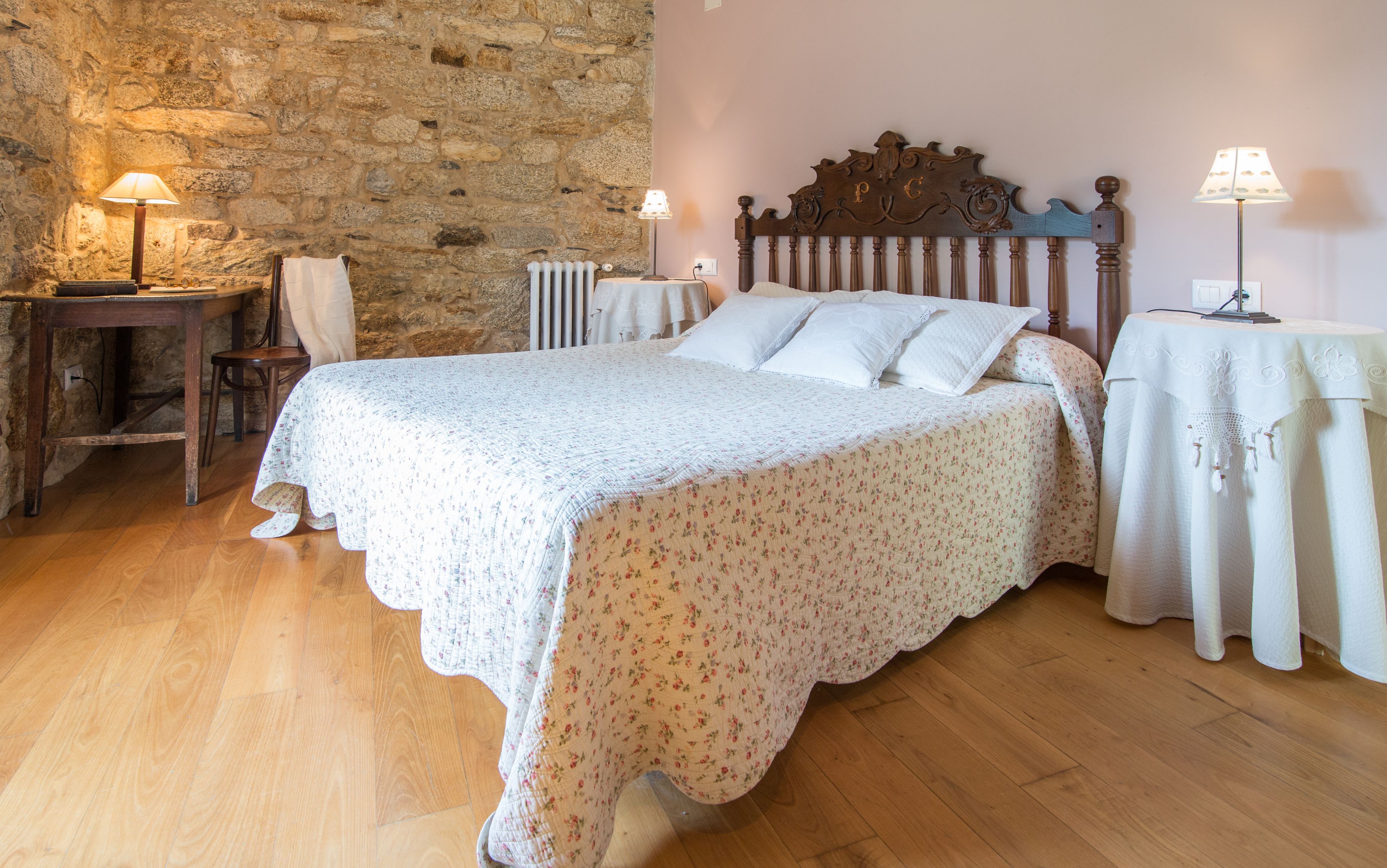 A bedroom with a stone wall, a wooden bed, two bedside tables with lamps, a chair, and a radiator.