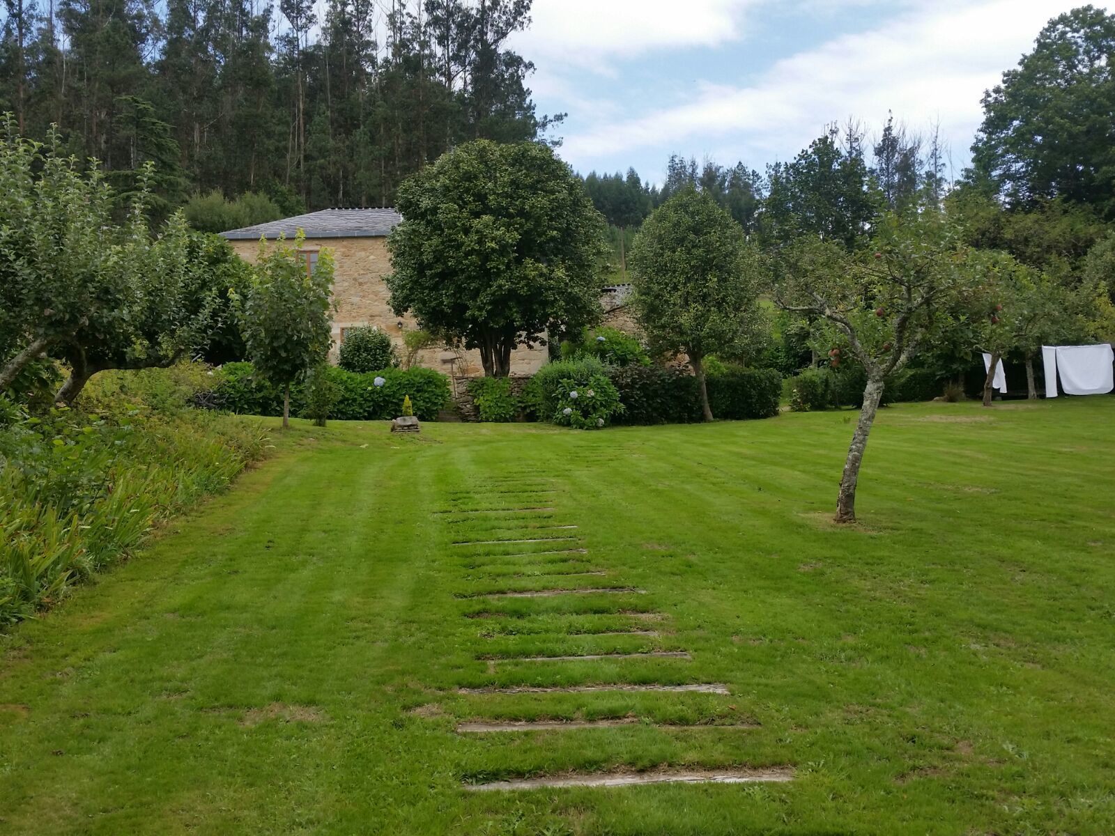 Een verzorgde tuin met een stenen gebouw, strak gemaaid gras en diverse bomen en struiken.