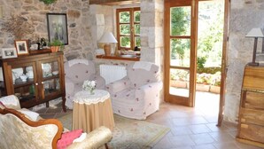 Una habitación rústica con paredes de piedra, un armario de madera, un sillón blanco, una mesa redonda y una ventana con vistas a la vegetación.