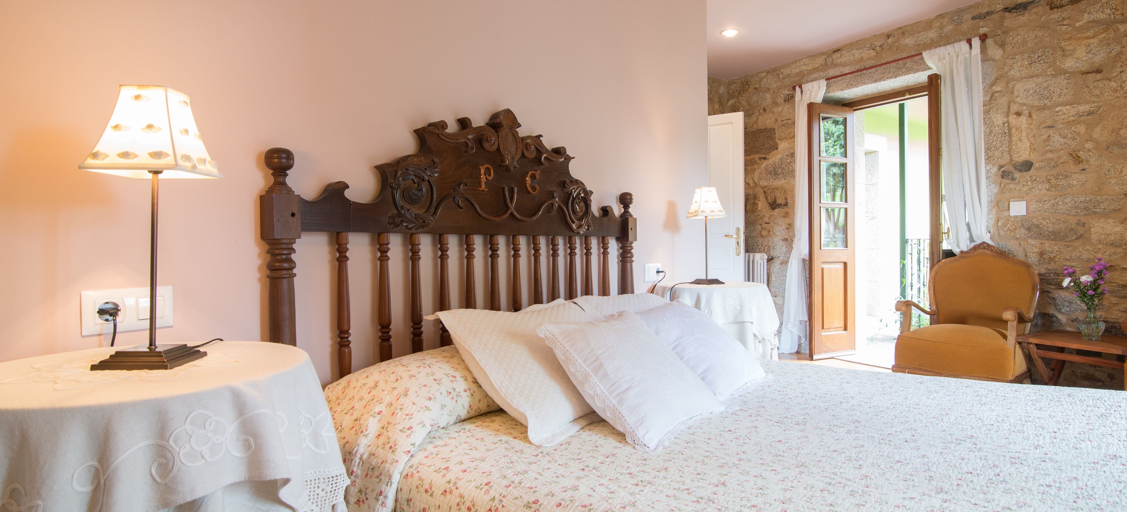A bedroom with a wooden headboard, a bed with floral bedding, two bedside tables with lamps, and a view of the outdoors through a door.
