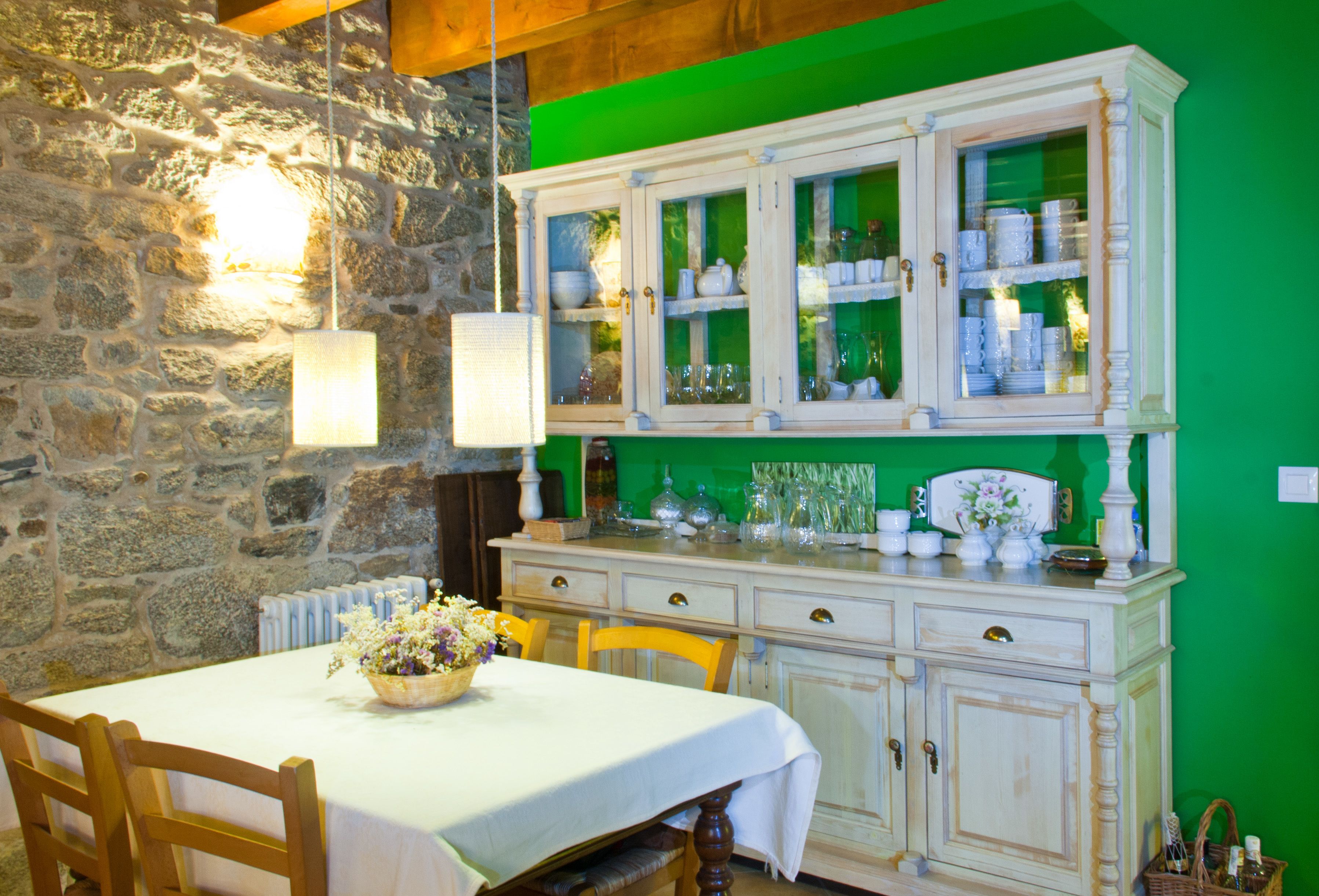 A dining area with a stone wall, a table with a white tablecloth, wooden chairs, and a cabinet with glass doors displaying dishes and glassware.