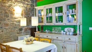 A dining area with a stone wall, a table with a white tablecloth, wooden chairs, and a cabinet with glass doors displaying dishes and glassware.