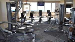 A gym with various exercise machines, a large window with a view of the ocean, and a "No Exit" sign.