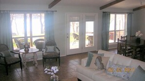 Ein geräumiges Wohnzimmer mit einem weißen Sofa, einem Esstisch und Stühlen. Große Fenster mit Vorhängen, ein Deckenventilator und eine Tür, die nach draußen führt.