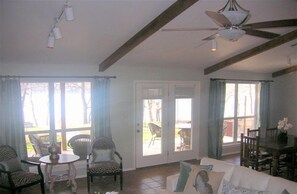 Ein geräumiges Wohnzimmer mit einem weißen Sofa, einem Esstisch und Stühlen. Große Fenster mit Vorhängen, ein Deckenventilator und eine Tür, die nach draußen führt.