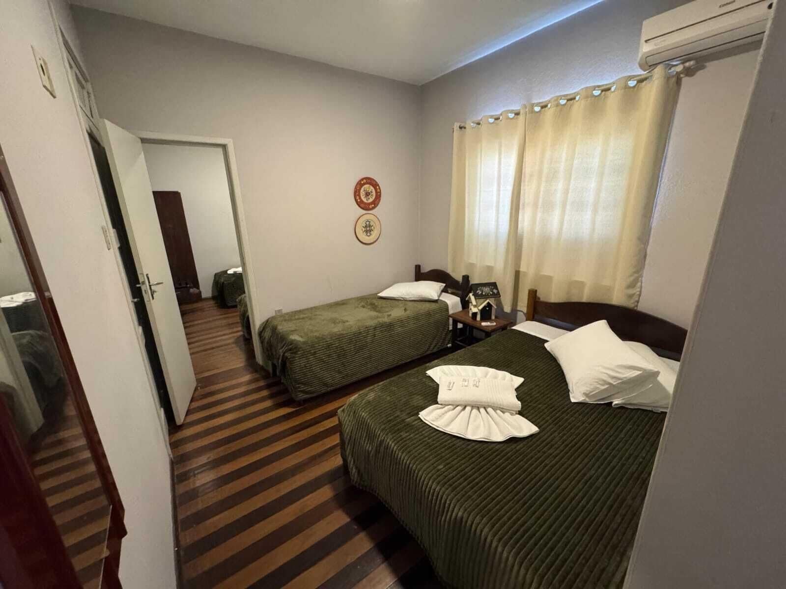 Quarto de hotel com duas camas de solteiro, piso de madeira, uma mesa pequena com abajur e ar-condicionado fixado na parede.