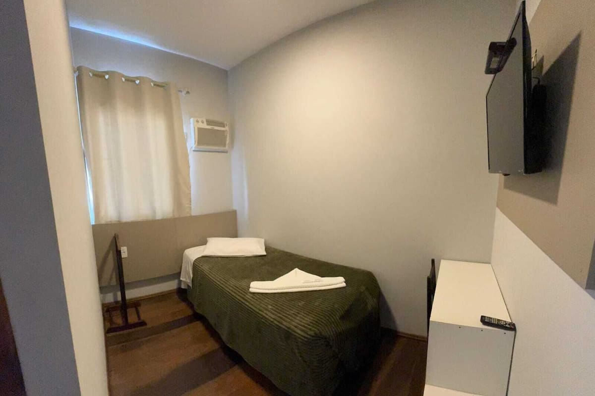 A small, single-bed room with a wooden floor, a flat-screen TV mounted on the wall, and a window with curtains.