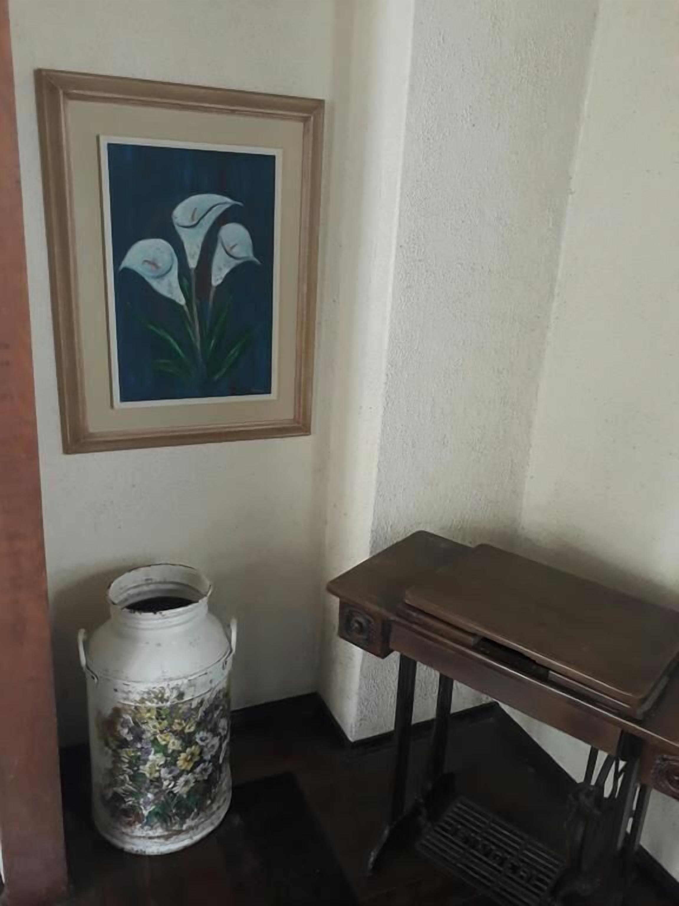 Eine Ecke mit einem gerahmten Blumenbild, einem weißen Keramiktopf mit floralen Verzierungen und einem kleinen Holztisch.