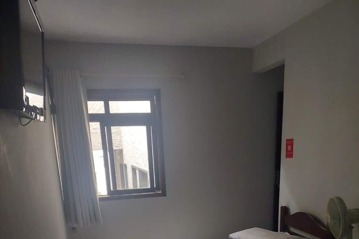 A hotel room with two beds, a window with curtains, a bedside table with a lamp, and a wall-mounted fan.
