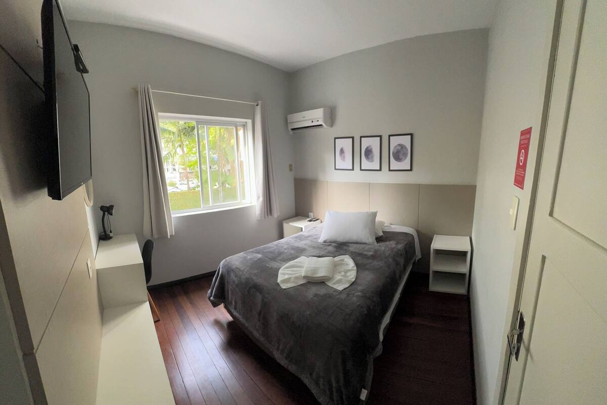 A hotel room with a bed, a desk, a chair, a TV, a window with curtains, an air conditioner, and framed pictures on the wall.