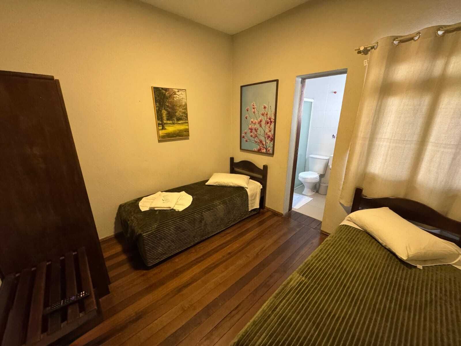 Una habitación pequeña y sencilla con una cama, una silla y un armario. Hay un cuadro en la pared y se ve un baño por una puerta abierta.