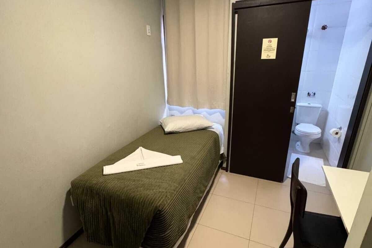 A small, single-bed room with a desk and chair, a window with curtains, and a door leading to a bathroom.