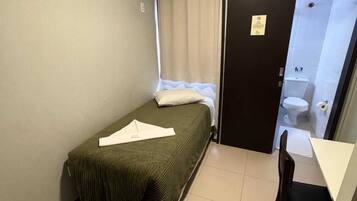 Una habitación pequeña con cama individual, un escritorio y una silla, una ventana con cortinas y una puerta que da al baño.