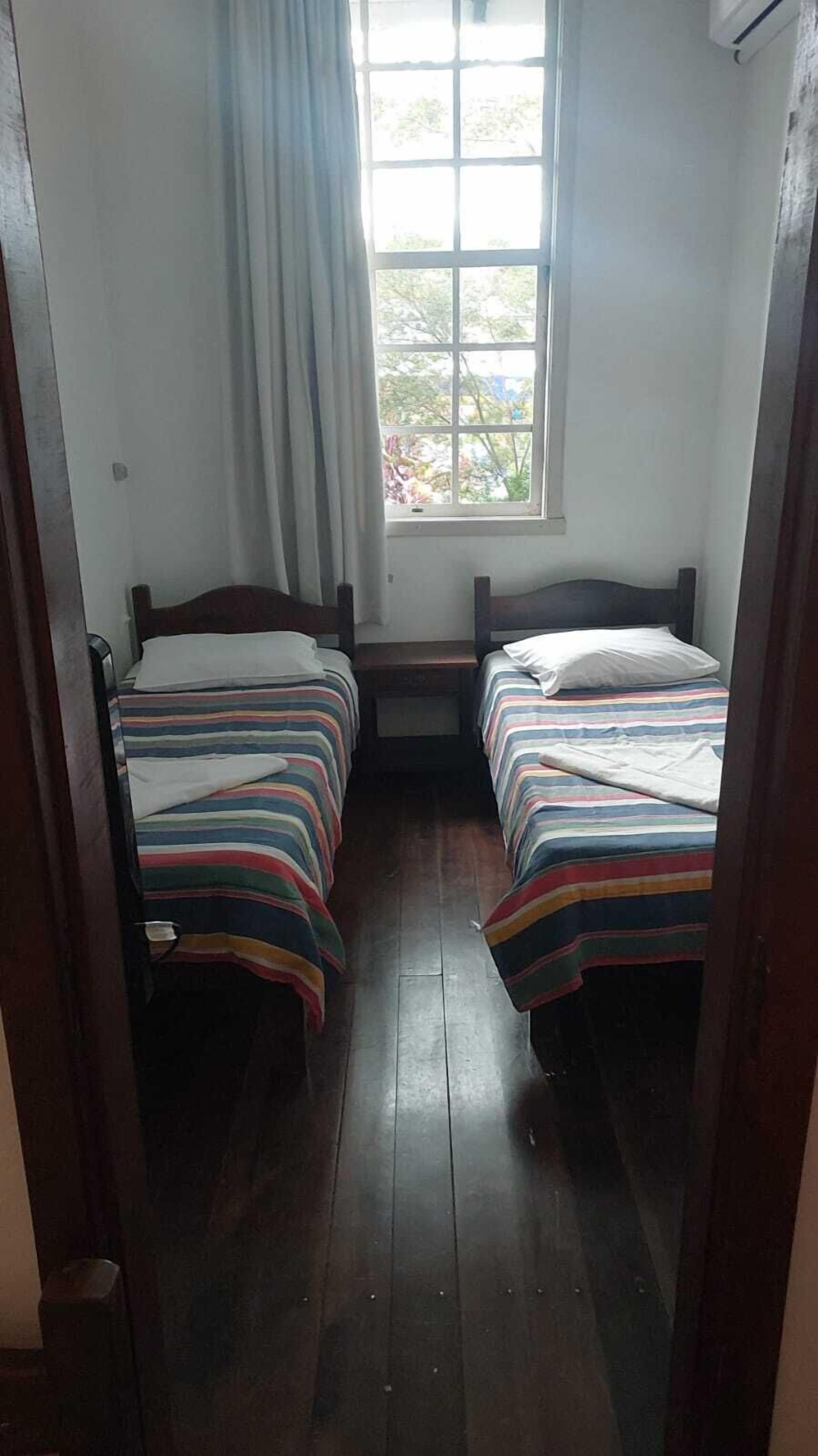 Una habitación pequeña y estrecha con dos camas de madera, una ventana con cortinas y piso de madera.