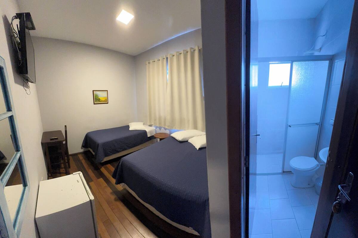 A hotel room with two single beds, a small table, a chair, a TV, and a bathroom with a toilet and shower.