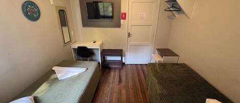 A small, compact room with two single beds, a desk with a chair, a TV mounted on the wall, and a door with a sign.