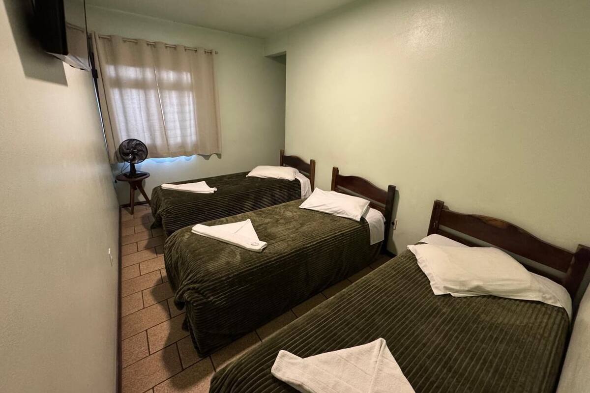 A room with three single beds, a small table with a fan, and a TV mounted on the wall.