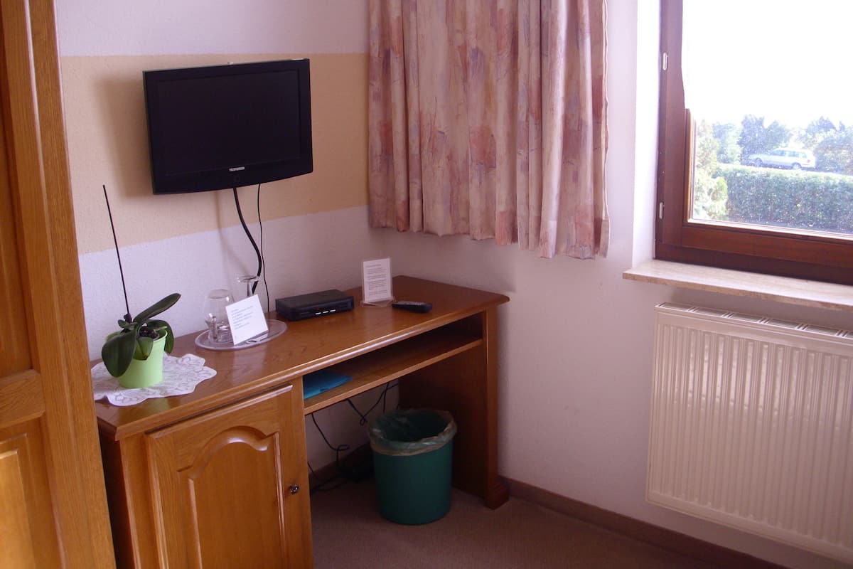 Quarto de hotel com secretária de madeira, televisão fixada na parede, janela com cortinas, radiador e lixeira.