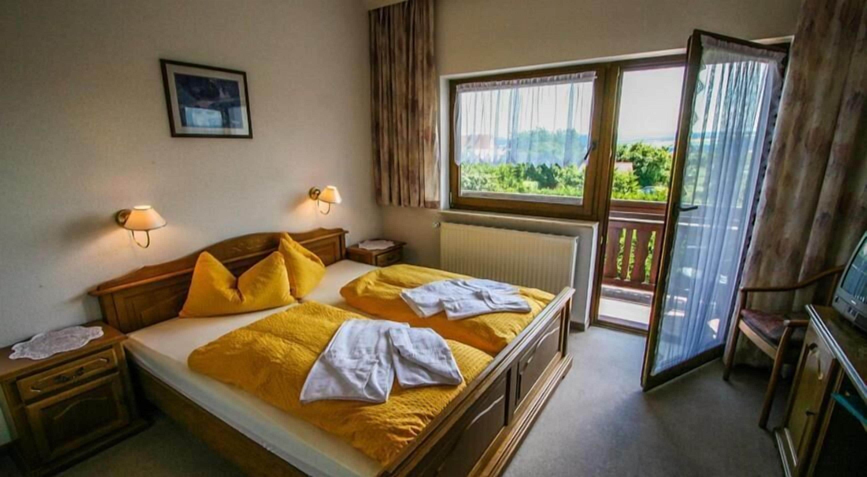 A bedroom with a bed, bedside tables, lamps, a window with curtains, and a view of greenery.