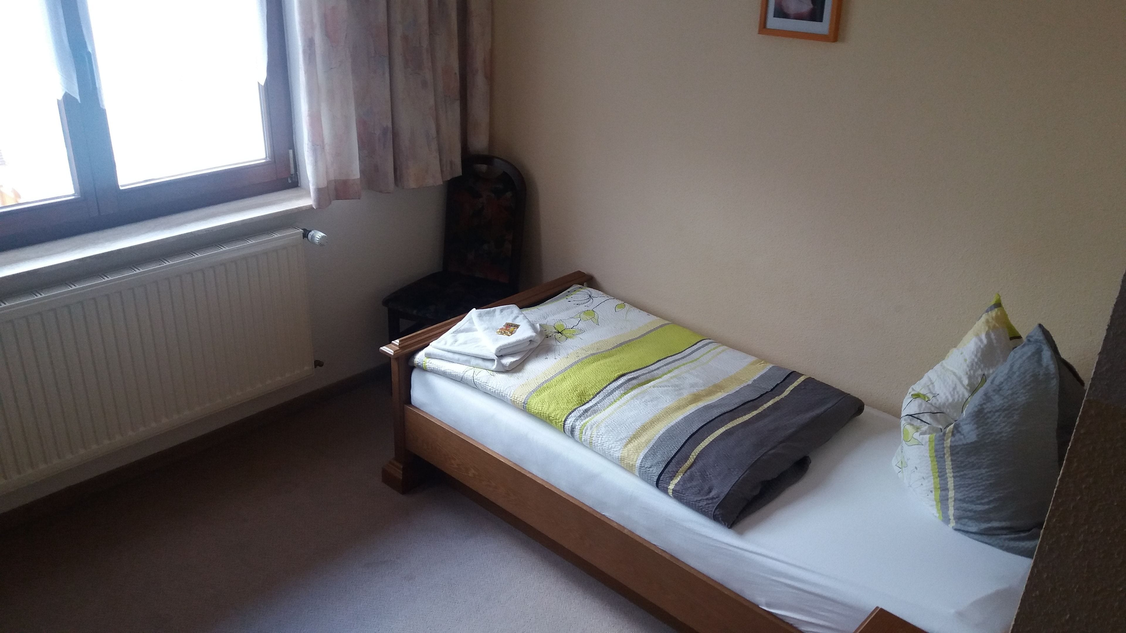 Une petite chambre avec un lit simple, un cadre en bois, un radiateur, une fenêtre avec des rideaux et une petite table de chevet.