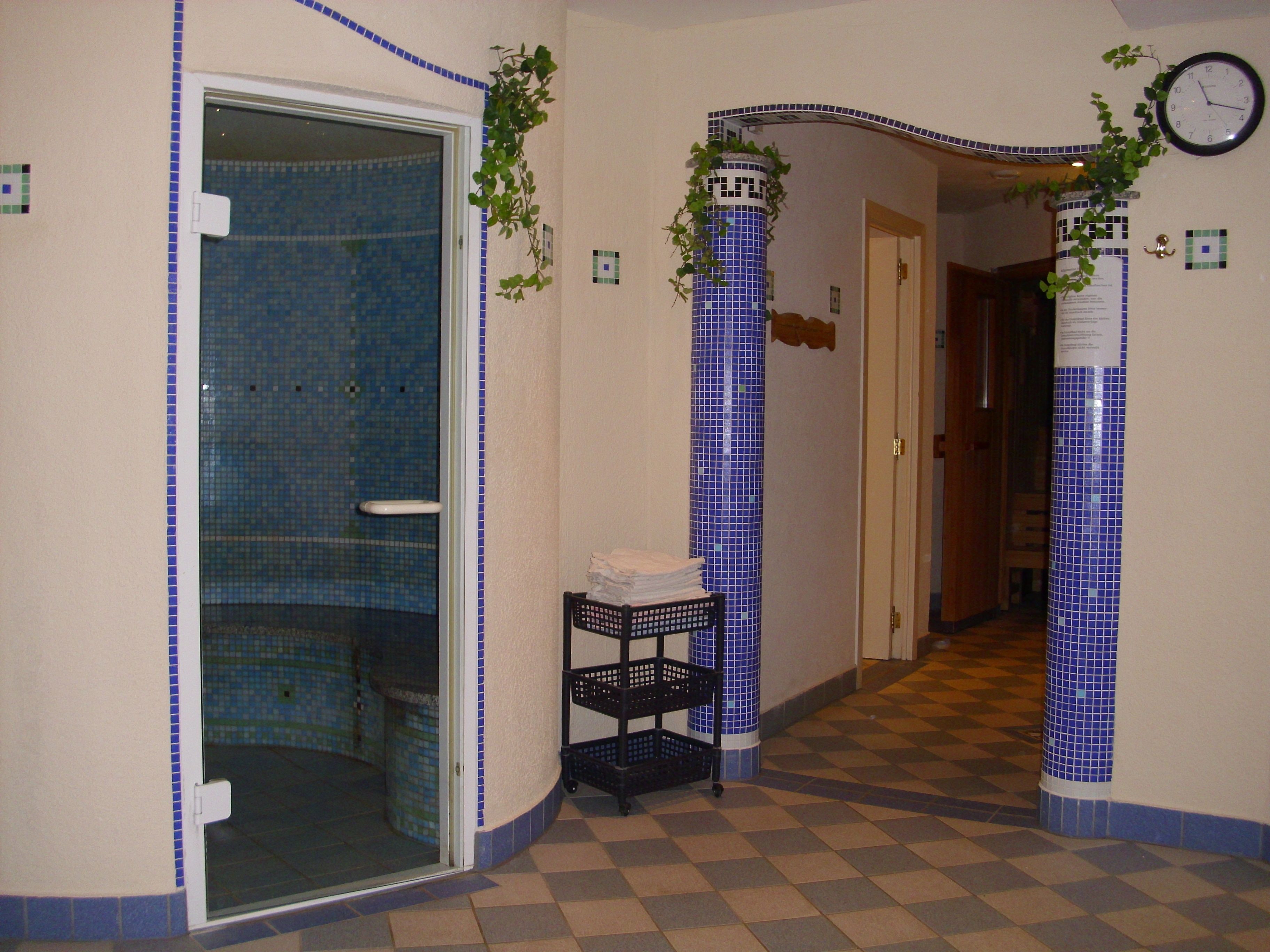 Uma casa de banho com porta de vidro, área de duche com azulejos, uma estante preta com toalhas e um relógio na parede.