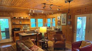 A cozy cabin interior with a wooden ceiling, a dining area, a kitchen, and a living room with a sofa and chairs.