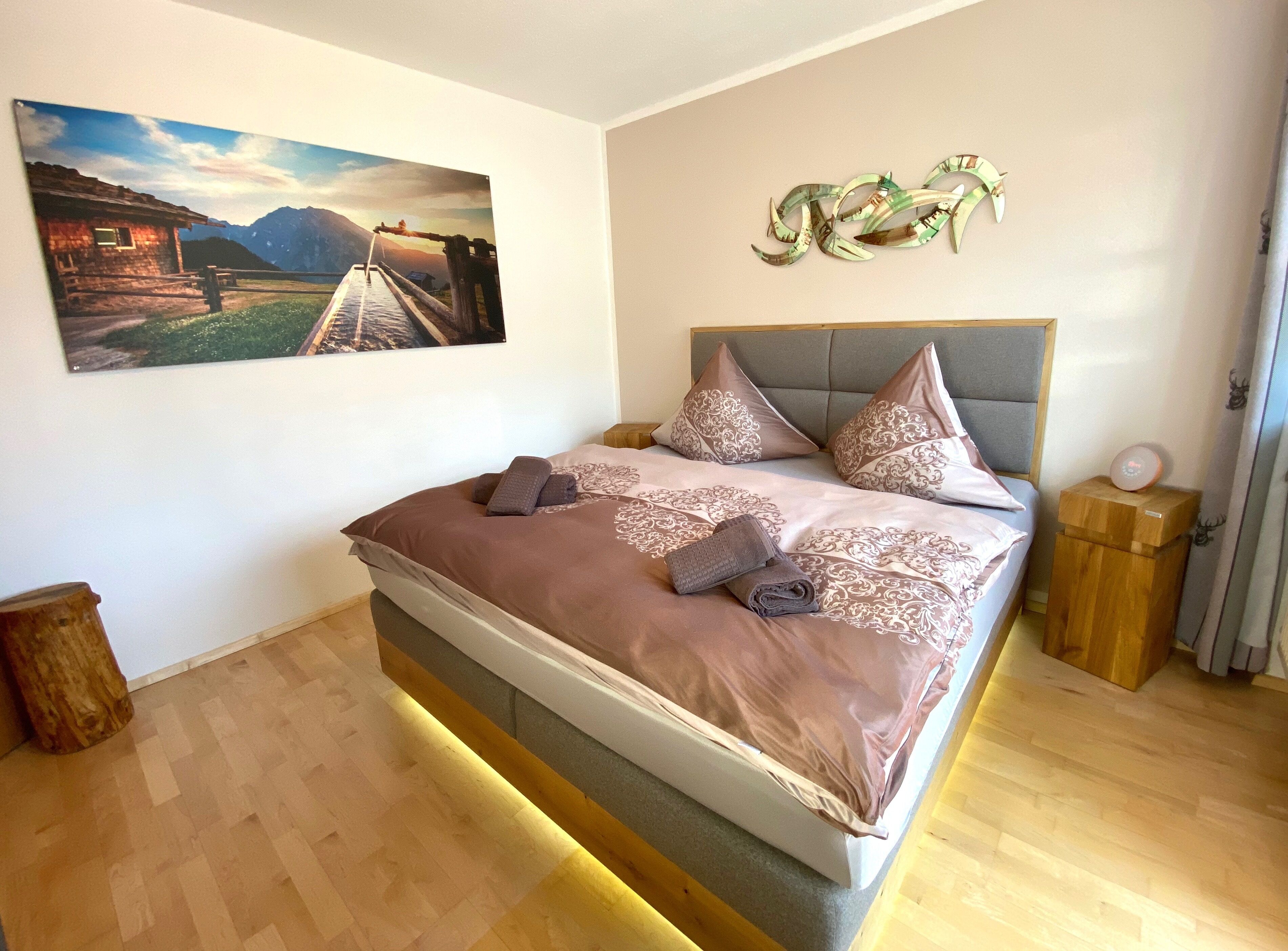 A neatly made bed with a headboard, a wooden nightstand, a decorative wall piece, and a framed picture on the wall.